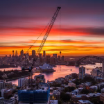 A striking, professional photo of a construction crane silhouetted against a dramatic sunset over the Sydney skyline, featuring the Sydney Opera House and Harbour Bridge. This image effectively visualizes the concept of a sunset clause in property development.
